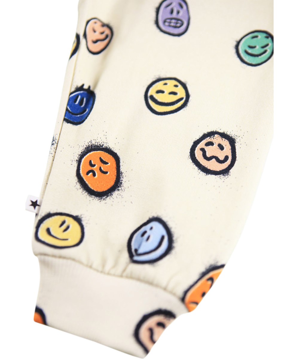 Simeon - Multi Moods - Weiche cremefarbene Hosen für die Kleinen aus Öko-Baumwolle mit einem Druck von bunten Gesichtern in verschiedenen Stimmungen.