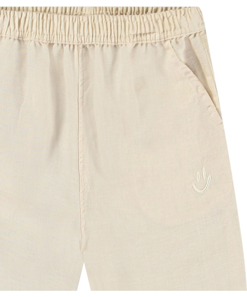 Sois - Summer Sand - Pants in organic cotton for baby. The pants are light beige with a small embroidered smiling face by the pocket.