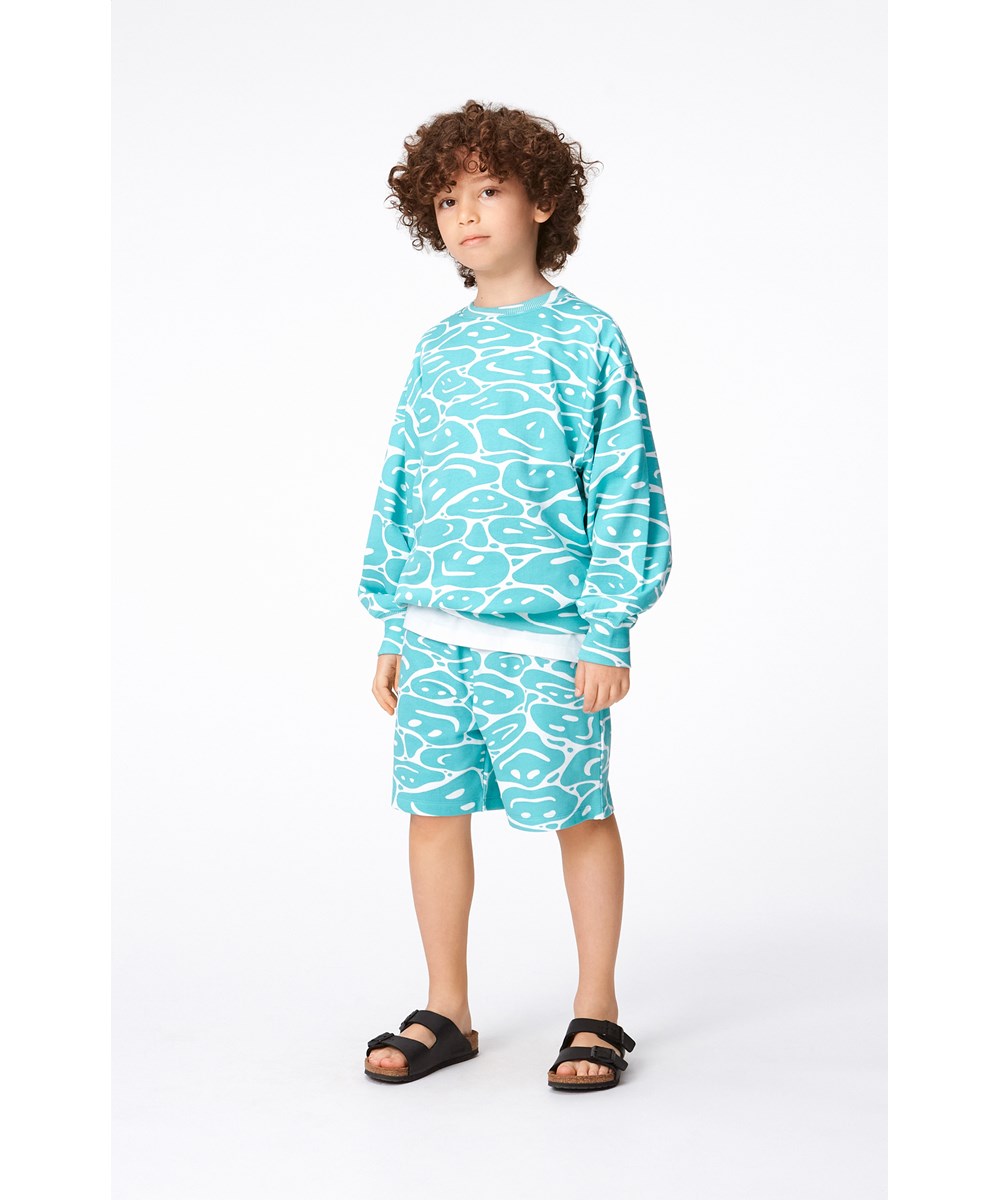 Monti - Waves Of Joy - Organic sweatshirt in turquoise and white in an all over print of smiling faces that are melting together 