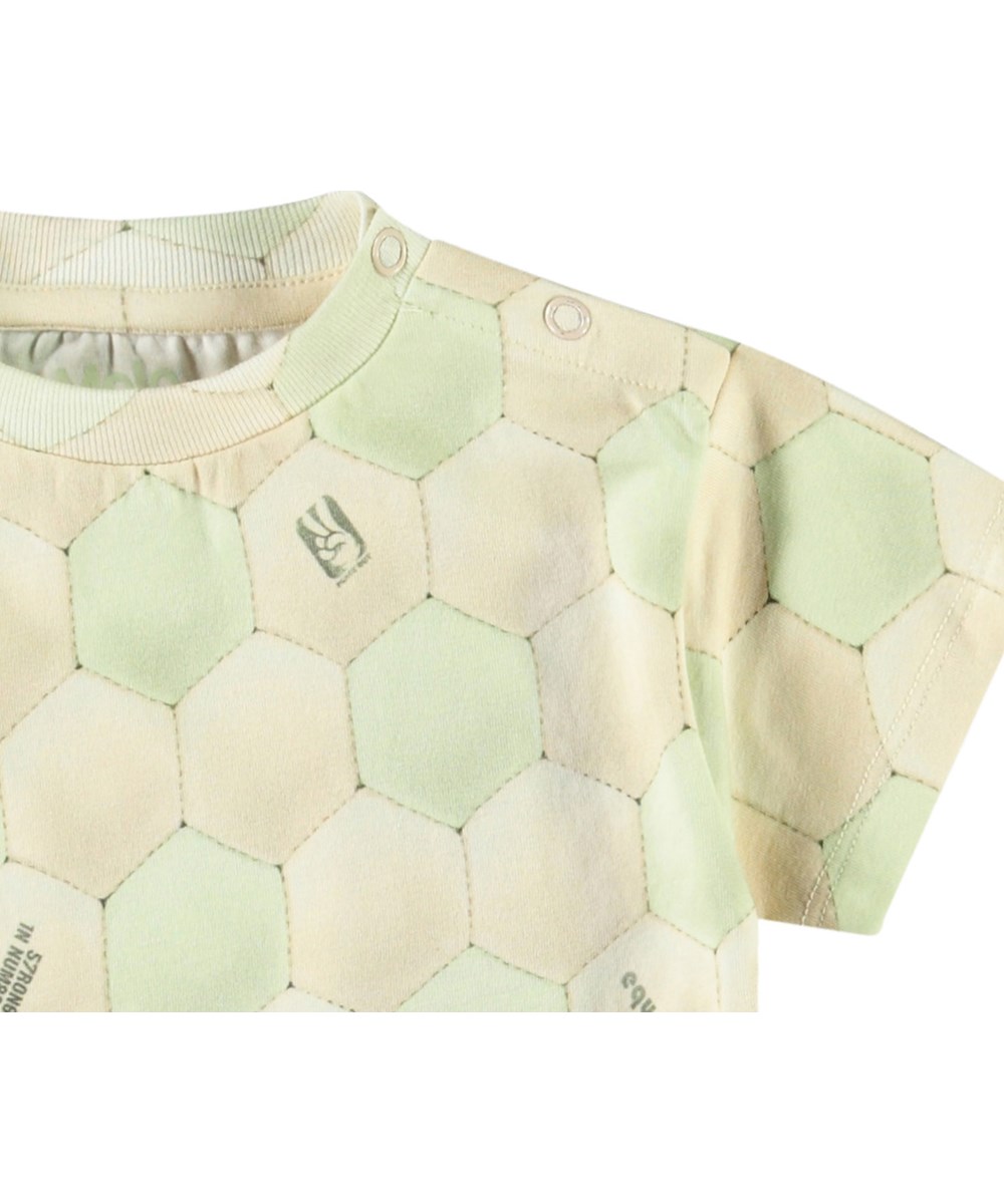 Enzo - Football Misty - Klassisches Öko-Baumwoll-T-Shirt für Babys. Das T-Shirt zeigt einen Druck einer Fußballoberfläche in Creme und Hellgrün.