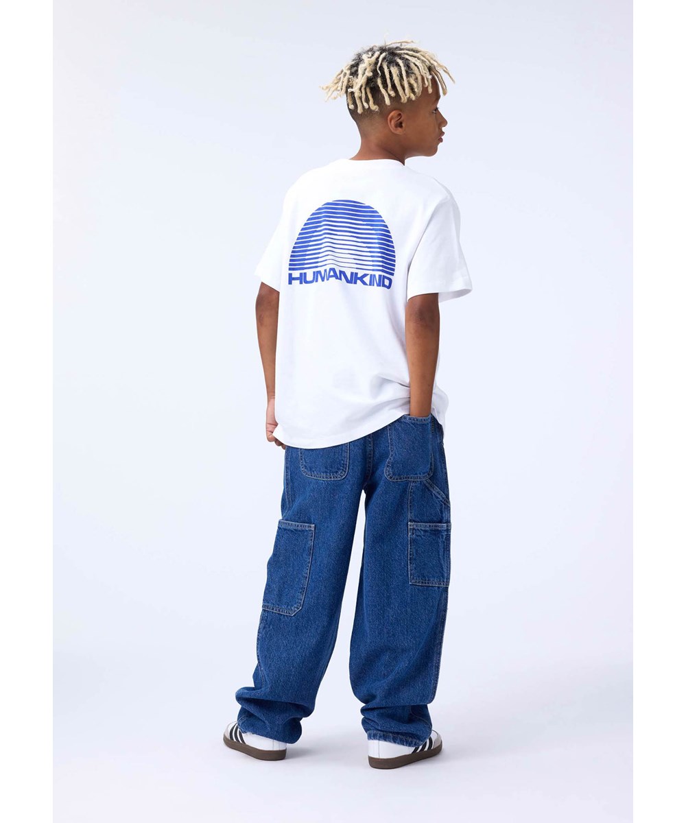 Riley - Humankind - Weißes T-Shirt für Kinder aus weicher Öko-Baumwolle mit kurzen Ärmeln, geripptem Ausschnitt und einem Aufdruck eines blauen Regenbogens mit dem Text ''Humankind''.