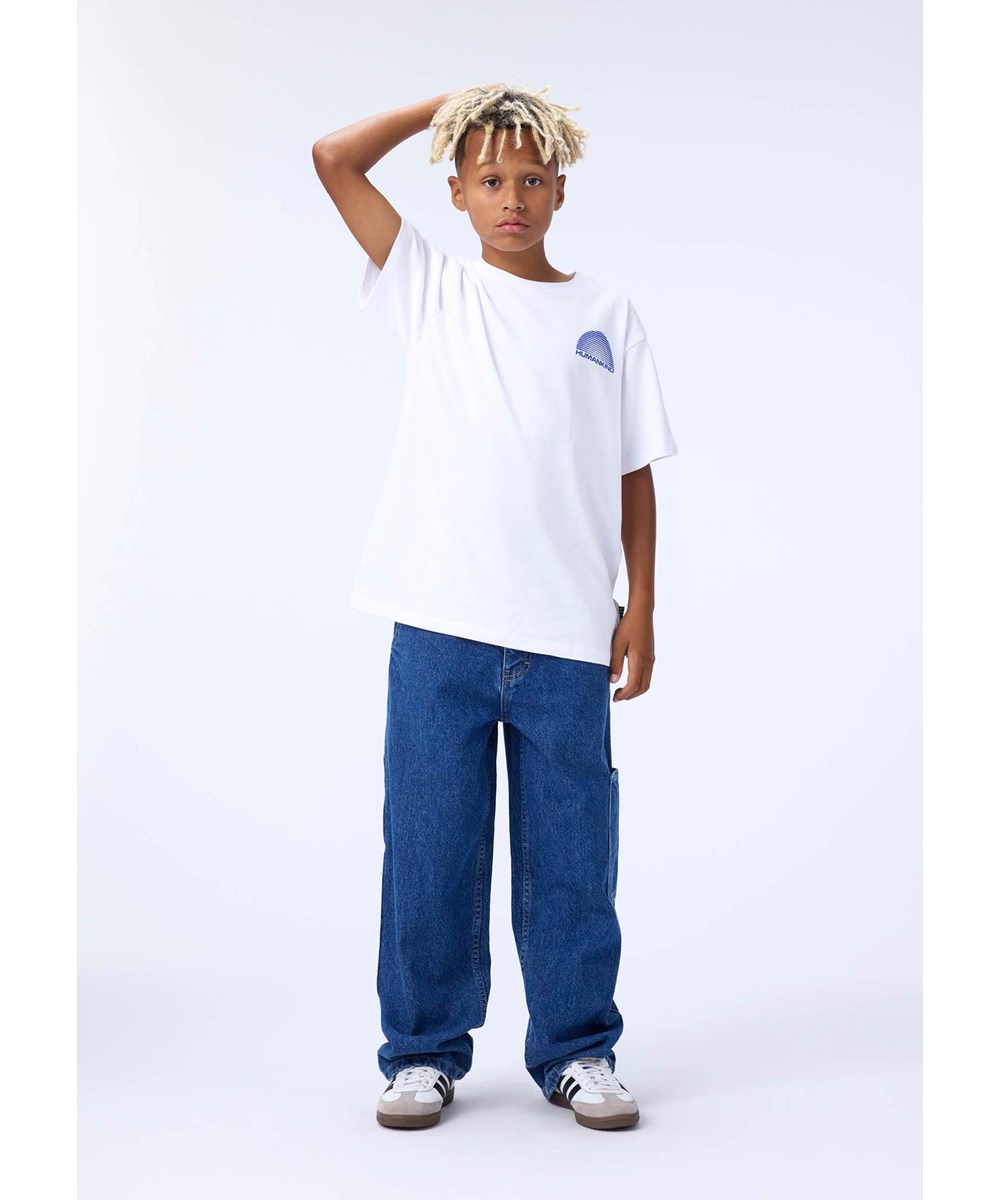 Riley - Humankind - Weißes T-Shirt für Kinder aus weicher Öko-Baumwolle mit kurzen Ärmeln, geripptem Ausschnitt und einem Aufdruck eines blauen Regenbogens mit dem Text ''Humankind''.