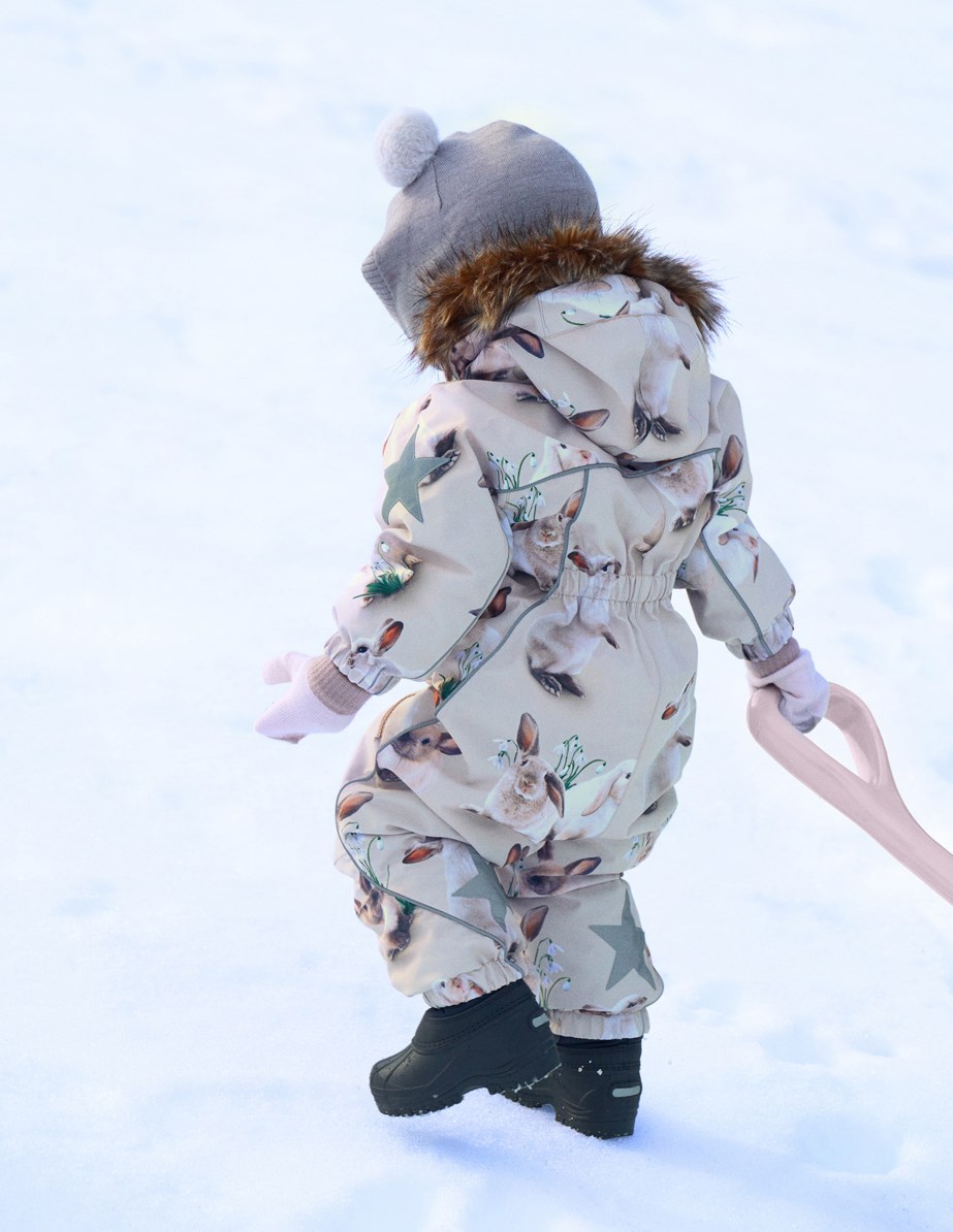Pyxis Fur - Winter Rabbits - Kinder-Schneeanzug aus recyceltem Polyester mit einem Druck von niedlichen Hasen zwischen Schneeglöckchen auf grauem Hintergrund.
