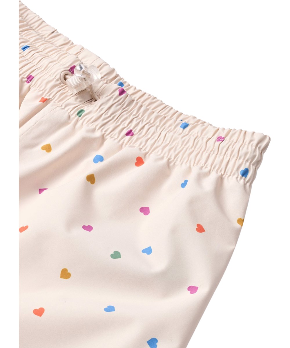 Waits - Confetti_Rainbow - Waterproof rain trousers in recycled polyester. The trousers are beige with a print of small hearts in various colours.