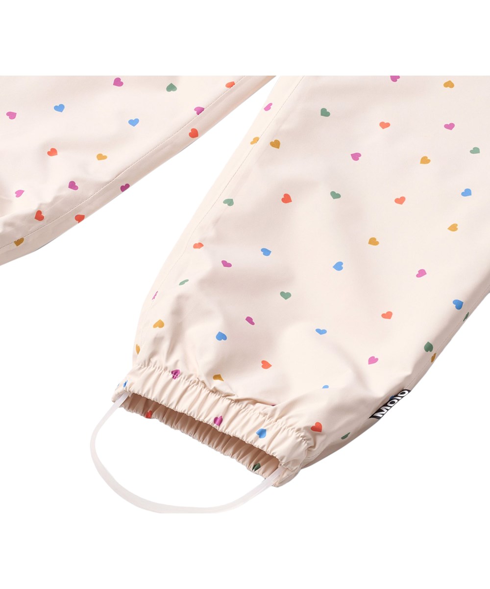 Waits - Confetti_Rainbow - Waterproof rain trousers in recycled polyester. The trousers are beige with a print of small hearts in various colours.