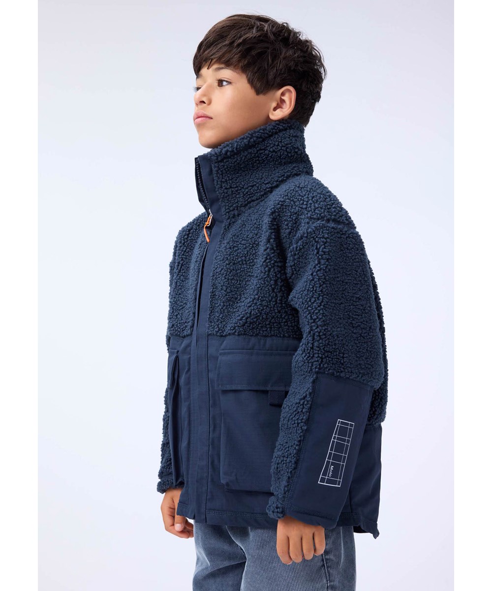 Utha - Night Navy - Mørkeblå fleecejakke til børn i genanvendt polyester med detaljer på ærmer, ryggen samt udvendige frontlommer i vind og vandafvisende slidstærkt nylon.