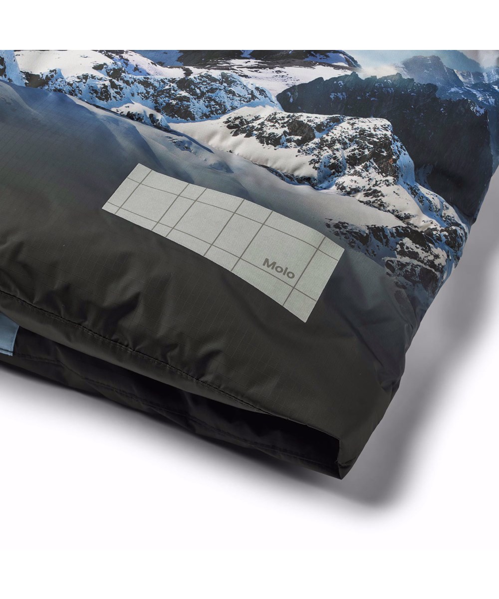 Hilo - High In Sky - Varm dynejakke til en kold vinter i genanvendt polyester, med et print af snedækket alper, solens skær og en klar blå himmel. Jakken har indbyggede vindfang i ærmerne og reflekser for øget synlighed i trafikken