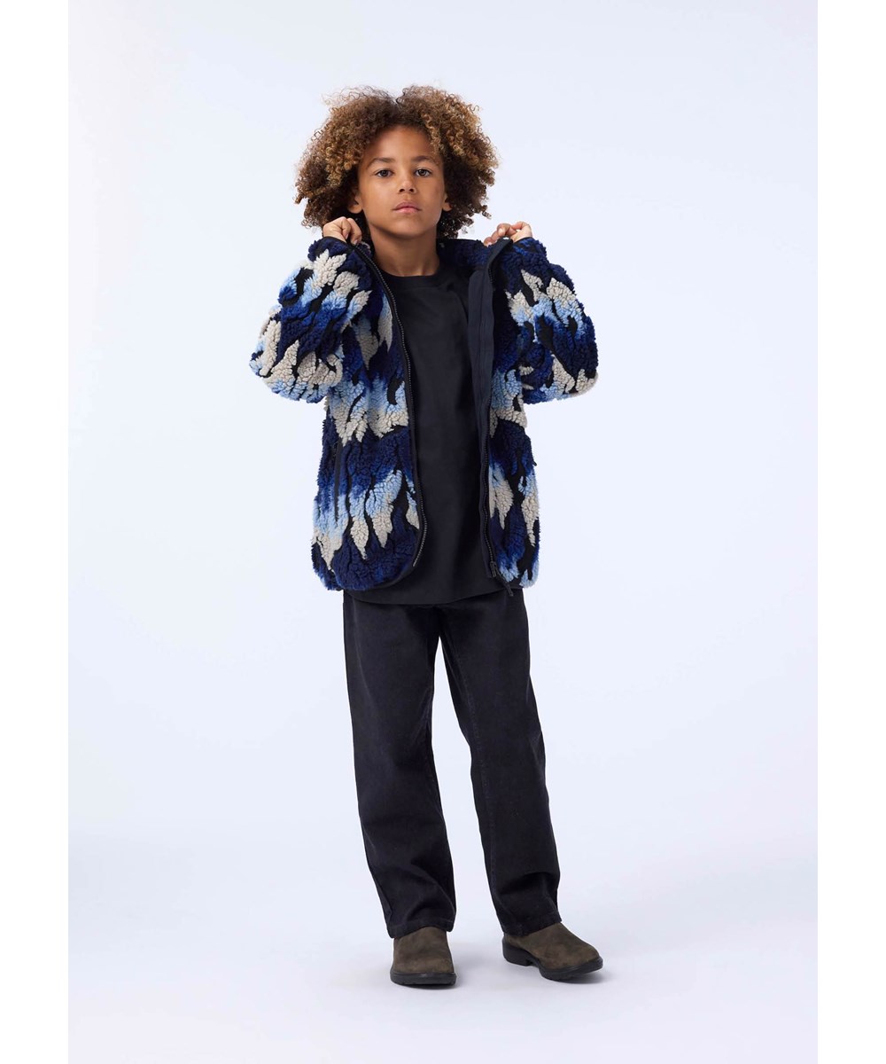 Unique - Ingenuity Cool - Mjuk fleecejacka för barn tillverkad av återvunnen polyester med matchande ribbade kanter och fodrad med mjuk bomull. Jackans mönster ser ut som lågor i blå och beige nyanser.